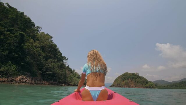 Sexy young woman in blue swimsuit swings feet in water on pink canoe on sea against hills backside view. Traveling to tropical countries. Attractive sportive girl is sailing on kayak in ocean.