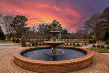 a circular water fountain in the garden surrounded by red brick, bare winter trees and lush green trees with red sky and powerful clouds at sunset at Memphis Botanic Garden in Memphis Tennessee USA