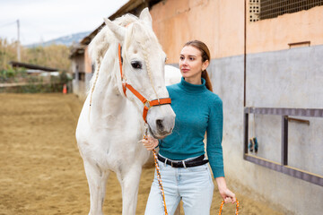 Confident young woman taking white horse to outdoor riding arena for training, leading by reins