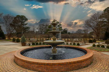 a circular water fountain in the garden surrounded by red brick, bare winter trees and lush green...