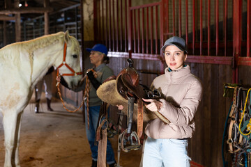 Young confident female stable keeper carrying horse saddle and harness for horseback ride..