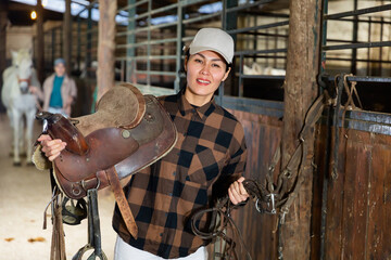 Positive woman carries harness for a horse in a stable