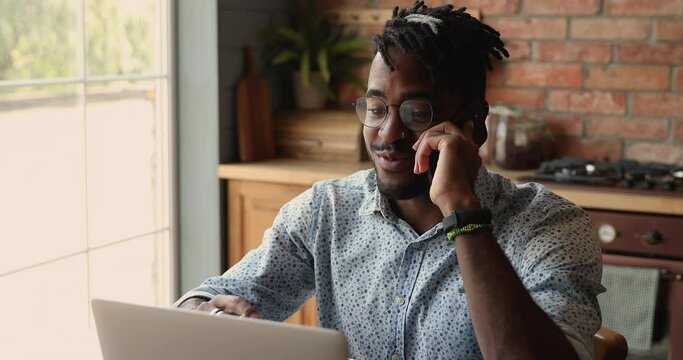 Optimistic young Black male freelance worker hold phone conversation before laptop screen share business success with partner colleague. Happy millennial male remote specialist consult client by phone
