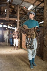 Portrait of young horsewoman walking in stable with saddle in hands, about to harness horse for...