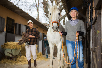 Skilled aged farmer woman leading white horse outdoors along stables in farm, holding by reins....