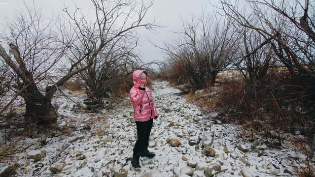 A young woman in a winter pink down jacket with a hood walks in the forest in winter. Beautiful frosty nature, dry forest in the snow. Dry mouth of the river, dark magical view.
