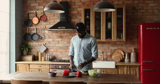 Inspired Afro American guy engaged in easy cooking vegetarian food cut vitamin salad of fresh vegetables at modern designed kitchen. Happy young male prepare simple tasty dietary breakfast on weekend