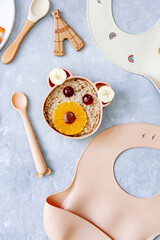 Funny bowl with oat porridge with bear faces made of fruits and berries on blue background. Food for kids idea, top view, copy space.
