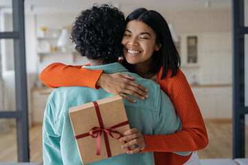 Beautiful smiling African American woman holding gift box. Happy birthday concept. Lovely multiracial couple hugging together at home 