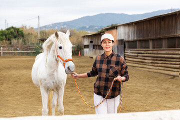 Asian woman rancher holding rein and leading horse outdoors.