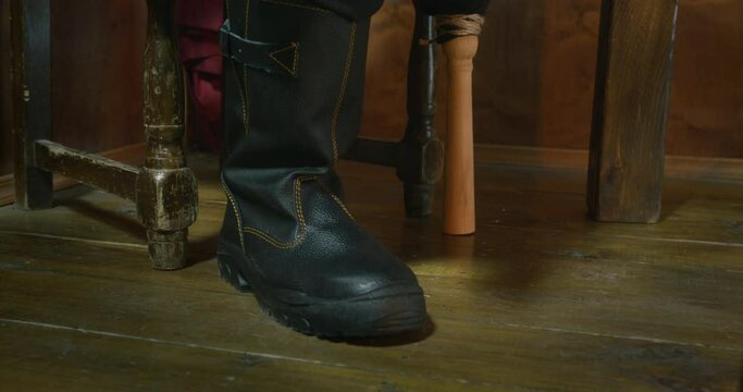 Pirate captain sits on old chair putting wooden leg prosthesis and right foot in black boot ON floor in ship cabin at voyage closeup