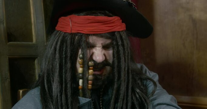 Crazy pirate captain with dark long braids and beard in cocked hat laughs sitting near windows IN cabin of ship close view