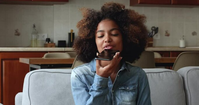 Beautiful 20s curly haired African teenage girl sits on sofa at home holding cellphone talks to friend on speaker phone, leaves audio message. Young gen use modern tech, remote communication concept