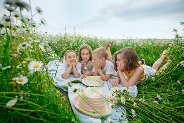 Obraz na płótnie Canvas A happy group of children lies in a field of camomiles at sunset in the summer. Boys and girls lie on a blanket in nature, a meadow with daisies. The concept of summer and childhood.