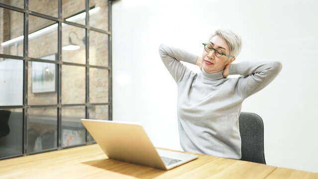 Office syndrome concept. Mature asian business woman wears glasses using laptop computer sit at workplace desk. She stretch her arm and relax neck.