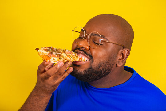 handsome afro brazilian man wearing glasses, blue shirt over yellow background. Eating pizza slice, cheese stretching, tasty, delicious.