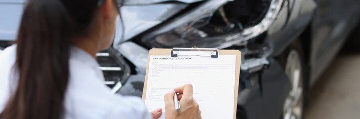 Woman agent filling out insurance claim form near wrecked car closeup