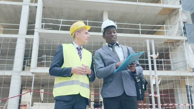 European foreman and African-american engineer in hardhats standing on building site and talking about documentation.