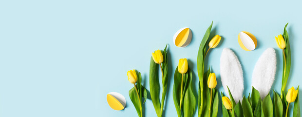 Yellow tulips, paper crafting eggs and white fluffy rabbit ears on blue background, easter and spring concept