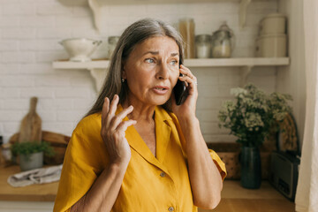 Elderly adorable Caucasian grandmother talking on phone with her grandson standing at kitchen having anxious and worried face expression, looking through window, waiting him for lunch - 487027572