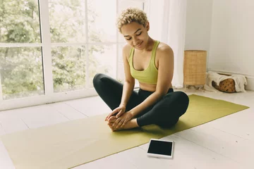 Fotobehang Light room with white panoramic window and African girl on the floor doing pilates in front looking with curious smile at pad screen with space for your advertisement. Body care, ethnicity concept © shurkin_son