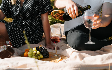 Love story, romantic picnic, valentine's day concept. Close-up of man pouring red wine into glass while sitting with woman on blanket with fruit outdoors. Young love couple on picnic in nature.