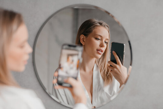 Pretty woman standing next to mirror in modern bathroom, holding smartphone and taking photo