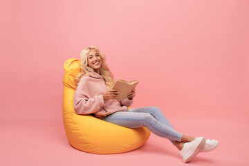 Cozy stay home activities. Relaxed lady reading her favorite book, sitting in beanbag chair on pink studio background