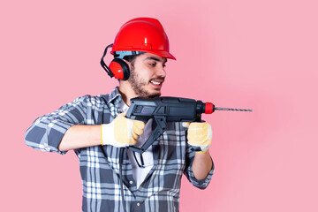 Young worker with a drill and helmet, on pink background.