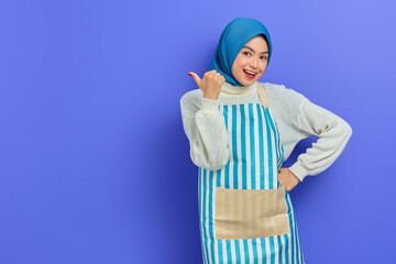 Portrait of smiling young housewife woman in hijab and apron looking at camera, pointing aside with finger isolated on purple background. People housewife muslim lifestyle concept