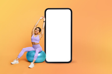 Fitness app. Athletic black lady training on fitness ball near big cellphone with blank screen, mockup
