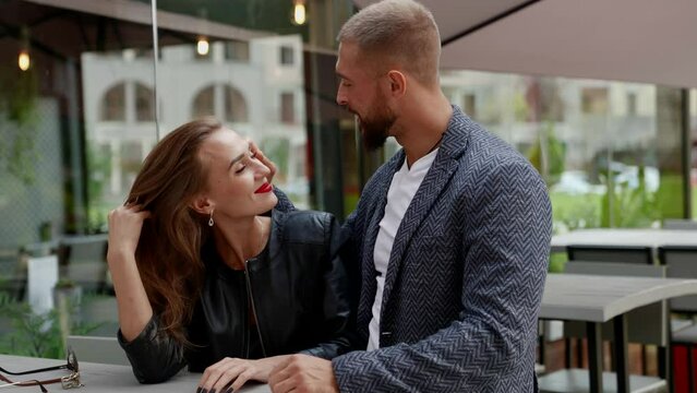 a man with a beard looks at a blonde with bright lipstick, then takes her hand at a cafe table on the street. the camera is moving
