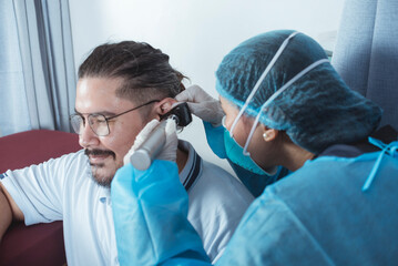 A doctor in full PPE gear checks the ear drum of a male patient with an otoscope, also known as an...