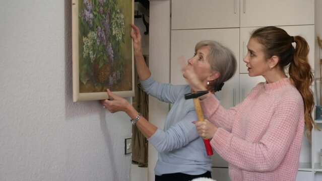 Adult caucasian woman helping her senior mother to put painting on wall. Daughter hammering nail, her mother standing beside with picture in hands. High quality 4k footage