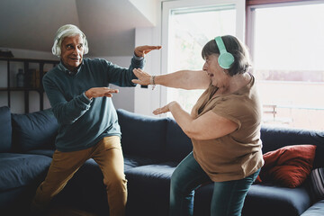 Nice active elderly couple enjoying a fancy dance in the living room wearing wireless headphones -...