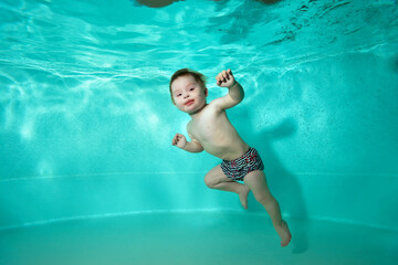 A child, a little boy, with Down syndrome swims underwater and poses in a pool with turquoise water. Horizontal orientation.