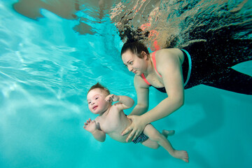 A happy mom and a child with Down syndrome swim together underwater in a turquoise water pool. The woman supports the baby with her hands and smiles. Portrait. Horizontal orientation.