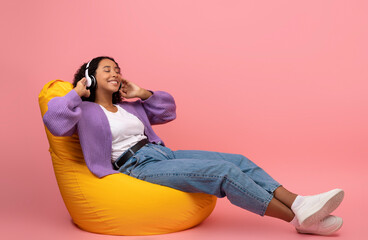 Full length of relaxed young black woman listening to music in headphones, resting in bean bag...