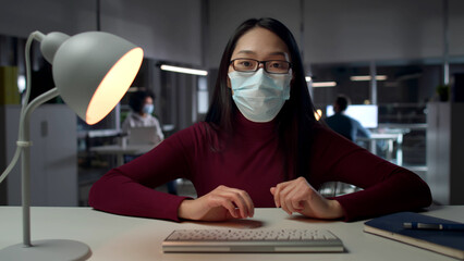 Female employee in safety mask sit at desk typing on computer keyboard