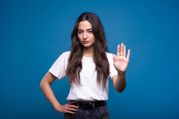 Attractive caucasian or arab brunette girl in a white t-shirt showing her palm as a stop gesture and frowning isolated in on a blue studio background.