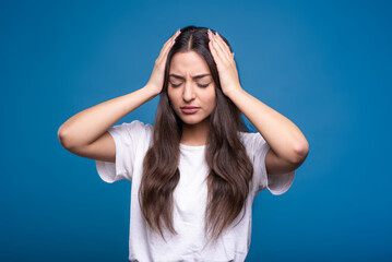 Attractive caucasian or arab brunette girl in a white t-shirt holding her hands behind her head because of a headache isolated on a blue studio background.