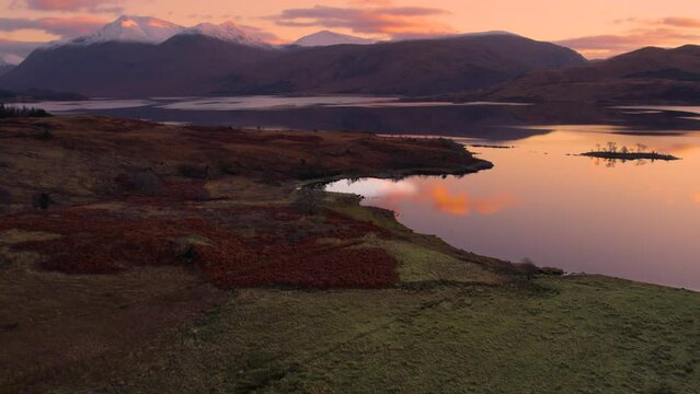 Aerial drone footage flying slowly away from Glen Etive and Loch Etive in Scotland during an orange and pink sunrise of pastel colours, with snow-capped mountains, trees and still reflective water.