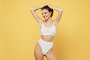 Fototapeta premium Smiling minded attractive young brunette woman 20s wearing white underwear with perfect fit body standing ties hair in ponytail look camera isolated on plain yellow color background studio portrait