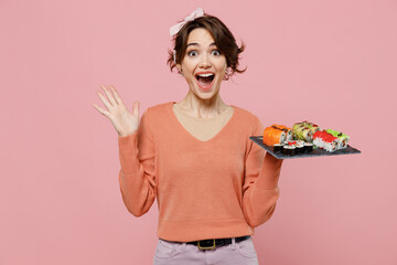 Young surprised shockedexcited amazed fun woman 20s in casual clothes hold makizushi sushi roll served on black plate traditional japanese food spread hands isolated on plain pastel pink background