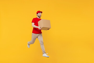 Full body fun side view delivery guy employee man in red cap T-shirt uniform workwear work as dealer courier jump high hold cardboard box run fast isolated on plain yellow background. Service concept.