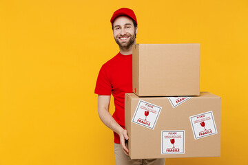 Professional happy delivery guy employee man in red cap T-shirt uniform workwear work as dealer courier hold cardboard box with fragile text isolated on plain yellow background studio Service concept - obrazy, fototapety, plakaty