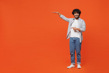 Fototapeta na wymiar Full size body length smiling happy young bearded Indian man 20s wear blue shirt pointing forefinger away down on workspace area copy space mock up isolated on plain orange background studio portrait