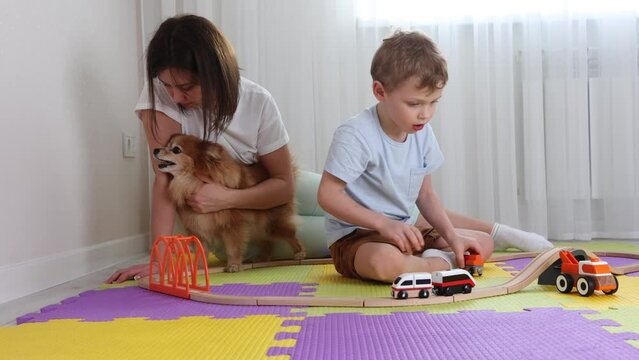 Happy mom, son and red dog spend time together playing a toy railroad in nursery on puzzle mats. Child spend the weekend with his mother, woman caresses a fluffy dog. Concept of free time and leisure
