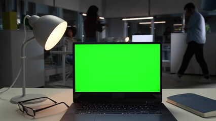 Laptop with green screen on desk in office with employees working on background