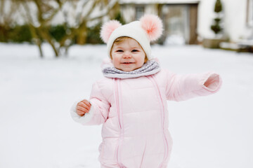 Happy little baby girl making first steps outdoors in winter through snow. Cute toddler learning walking. Child having fun on cold snowy day. Wearing warm baby pink clothes snowsuit and bobbles hat.